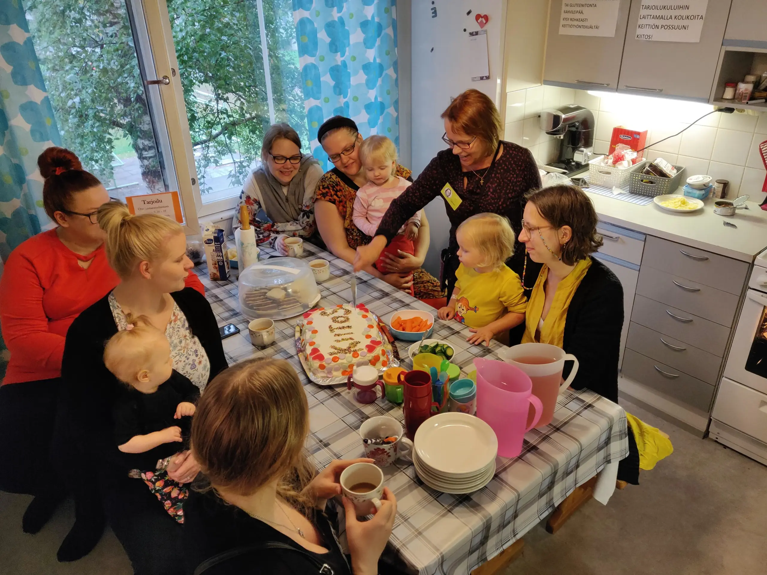 Iloiset aikuiset ja pienet kävijät Lähiötalon vanhan tilan keittiössä katsomassa, kun kohtaamispaikan perustaja Kirsti Kurki leikkaa kakkua jossa lukee Kiitos.