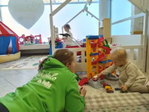 Lähiötalon työntekijä vihreässä hupparissa leikkimässä autoilla ja muovisella parkkihallilla pienen taaperon kanssa kohtaamispaikan lattialla Rantakylässä