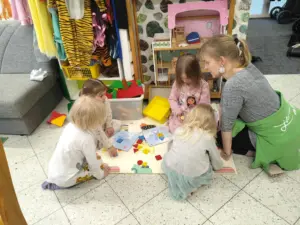 Lähiötalon johtaja Marika leikkimässä kohtaamispaikan lattialla neljän pienen lapsen kanssa rakennuspalikoilla. Taustalla näkyy myös nukketalo ja rooli-asut.